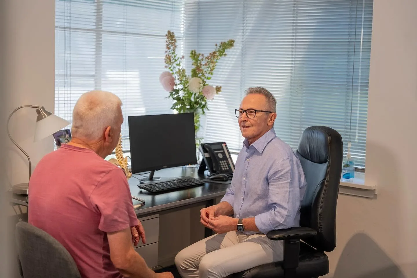 Dr Mark Navin consulting with a patient in the clinic's sunlit treatment room — spine model and monitor on the desk